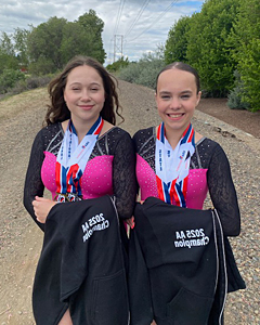 Two young gymnasts in pink and black leotards posing together with medals and black 2025 AA Champion jackets on a gravel path.